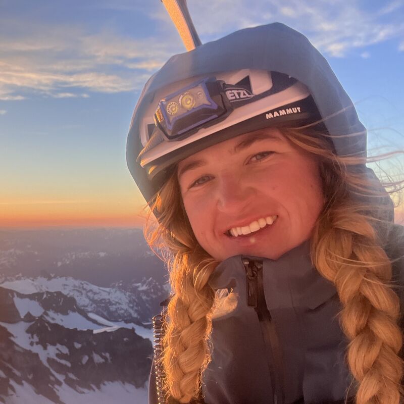 The image shows a woman with blonde braids wearing a helmet and a headlamp, likely on a mountain. She is smiling and appears to be at a high altitude, with snow-capped mountains visible in the background. The sky is bright with the colors of either sunrise or sunset, suggesting she may be mountaineering or engaging in a similar outdoor activity.